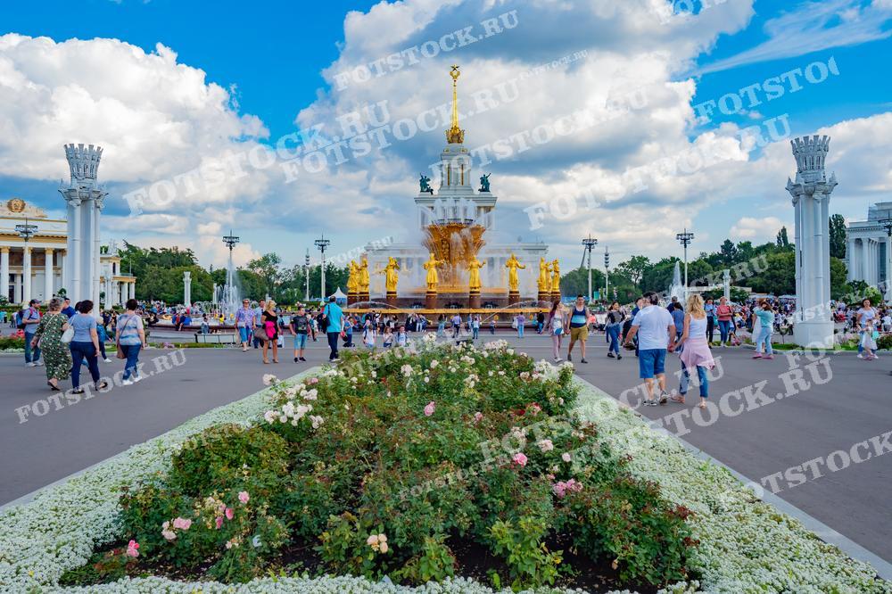 На фотографии изображен фонтан "Дружба народов" на ВДНХ в Москве. Вокруг фонтана гуляют люди, наслаждаясь летним днем. ВДНХ — это выставочный комплекс, где проходят различные мероприятия и выставки.