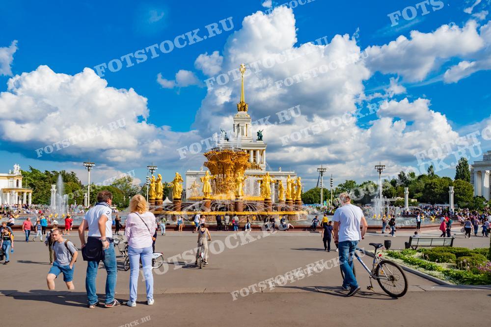 На фотографии изображен фонтан "Дружба народов" на ВДНХ в Москве в солнечный летний день. Вокруг фонтана гуляют люди, видны павильоны выставки. Фото можно использовать для иллюстраций, рекламы и туристических материалов.