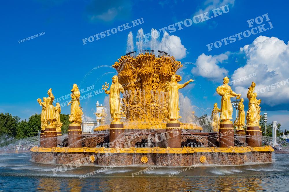 На фотографии изображен фонтан "Дружба народов" на ВДНХ в Москве в солнечный летний день. Золотые фигуры фонтана окружают центральную композицию. Фото можно использовать для иллюстраций, рекламы и туристических материалов.