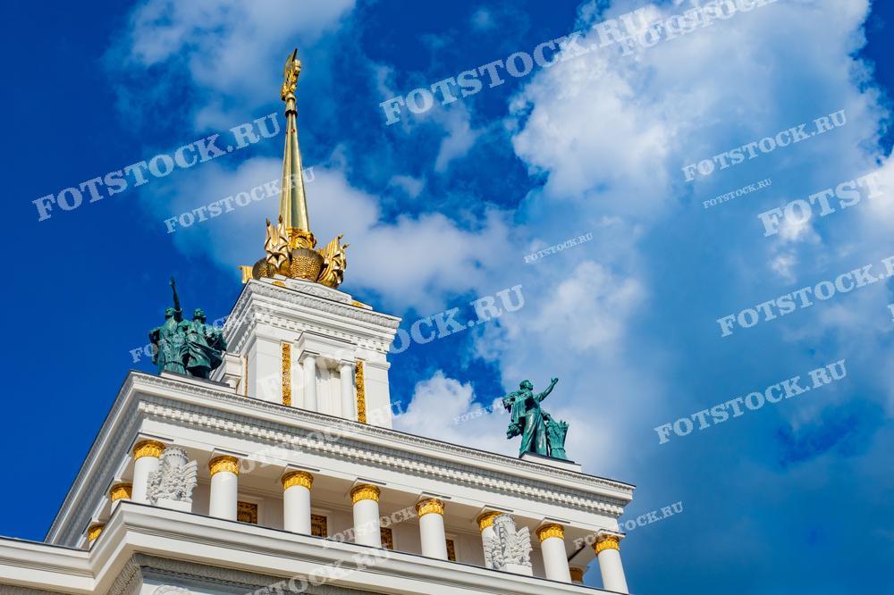 На фотографии изображен верхний фрагмент главного павильона ВДНХ с золотым шпилем и скульптурной композицией. Ясное синее небо подчеркивает архитектурные детали здания.
