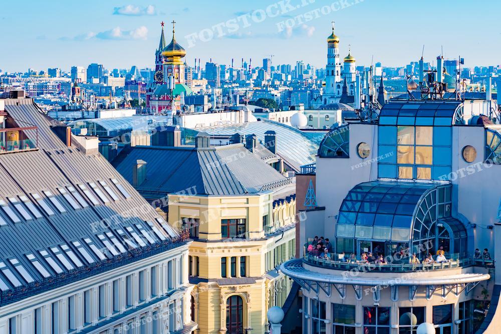 На фотографии изображена панорама Москвы, снятая с высоты. На переднем плане видны крыши современных зданий, а вдали можно увидеть колокольню Ивана Великого, Спасскую башню и другие исторические постройки.