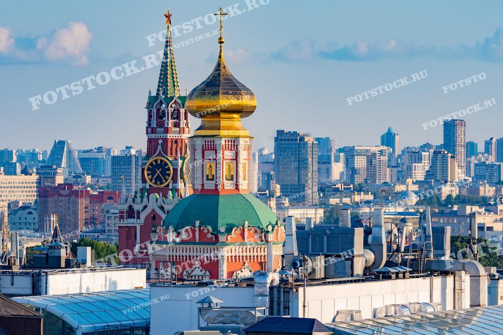 На переднем плане видна знаменитая Спасская башня Кремля с часами, символом Москвы и России, а также золотой купол храма на Красной площади. Эти исторические постройки находятся в центре Москвы и окружают Красную площадь. На заднем плане видны современные многоэтажные здания, подчеркивающие контраст между старинной архитектурой Кремля и динамично развивающимся городом. Снимок сделан в светлое время дня, позволяя насладиться яркими деталями архитектуры.