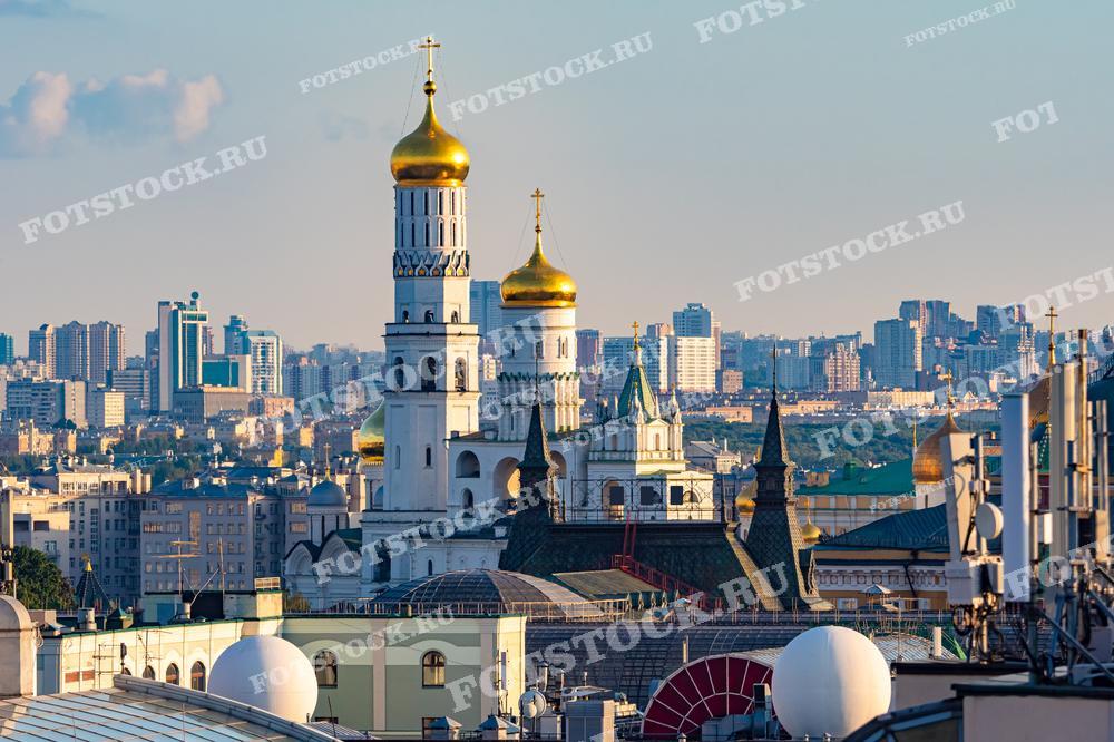 На фотографии представлен вид на колокольню Ивана Великого в Московском Кремле, окружённую другими историческими зданиями, на фоне современных высоток. Это сочетание старинной архитектуры и новых городских застроек иллюстрирует контраст между историческим наследием и современным развитием города. Фотография может использоваться для образовательных целей, в туристических публикациях, в культурно-исторических исследованиях, а также в прессе для иллюстрации статей о Москве и её архитектуре.