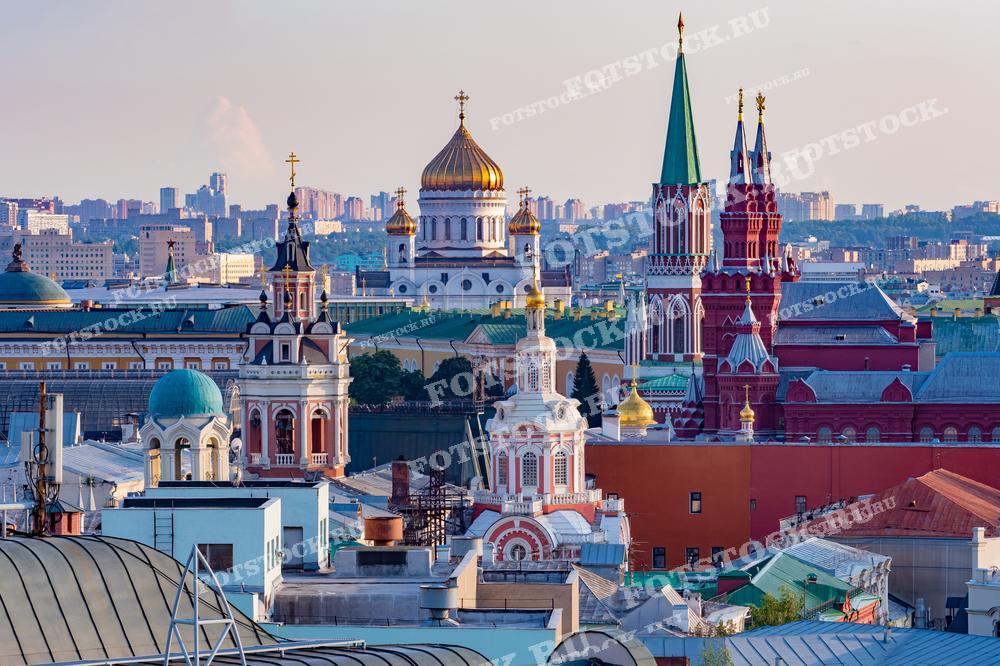 На фотографии изображена панорама Москвы с высоты, где видны храмы и башни. На переднем плане выделяются Храм Христа Спасителя и купола кремлевских церквей, создавая впечатляющий городской пейзаж.