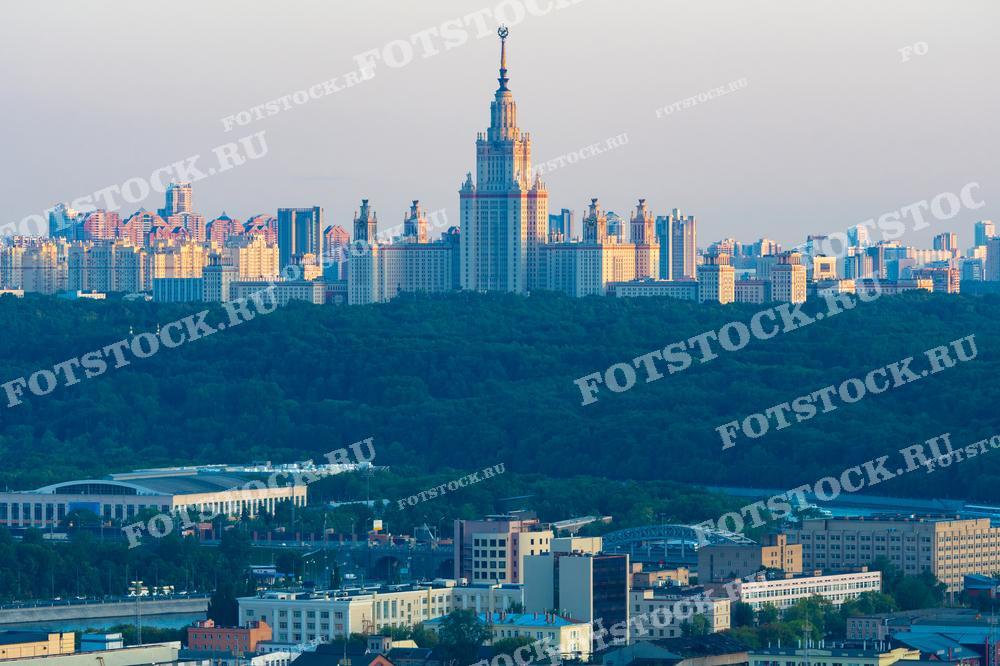 На фотографии изображена панорама Москвы с высоты, с видом на главное здание Московского государственного университета (МГУ) на Воробьевых горах. Вечернее солнце освещает городские кварталы, создавая теплую атмосферу.