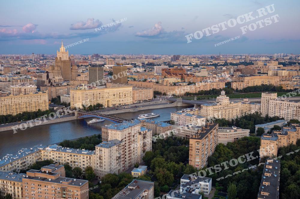 На фотографии изображён вечерний вид на Москву с высоты птичьего полёта. Особенно выделяется здание Министерства иностранных дел России, одно из высотных зданий, построенных в стиле сталинского ампира. На переднем плане видны жилые районы с характерными для советской эпохи многоэтажками, а также мост через Москву-реку, по которому проходит активное движение транспорта. Эта фотография может быть использована для иллюстрации статей о Москве, в туристических проспектах, исследованиях градостроительства и истории архитектуры.
