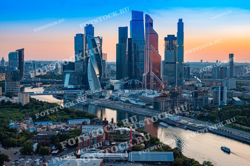 Фотография показывает панорамный вид на Москва-Сити, снятый с дрона. Высотные здания делового центра отражаются в реке Москва на фоне закатного неба.