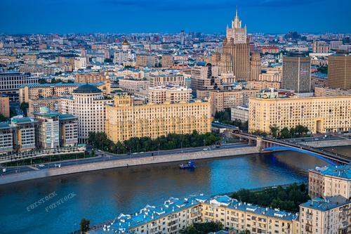 Фотография демонстрирует вечерний вид на Москву, охваченную тёплым светом закатного солнца. Здания с историческим фасадом в центре города гармонично сочетаются с более современными постройками вдалеке. Москва-река, пересекаемая мостом, отражает водную гладь небесного цвета, а золотой оттенок заката мягко ложится на сталинскую высотку, создавая пейзаж, наполненный спокойствием и величием столицы.