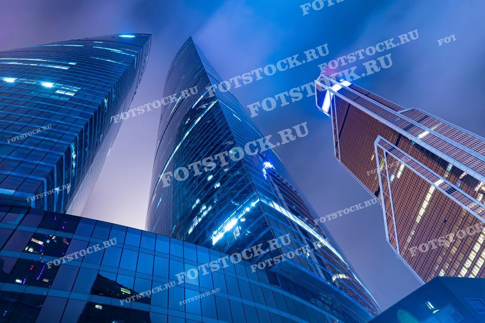 На фотографии запечатлены небоскрёбы Москва-Сити в вечернее время. Изображение демонстрирует впечатляющую игру света и архитектурные формы современных высотных зданий, которые стремятся в небо, создавая футуристический вид города. Эта фотография идеально подходит для использования в публикациях о современной архитектуре, статьях о городском дизайне, в презентациях бизнес-проектов, а также в материалах, посвящённых ночной жизни и культуре мегаполисов.