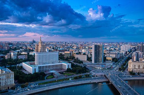 На фотографии запечатлен захватывающий вид на Москву в сумеречное время. Мощные облака нависают над городом, создавая драматичный фон для сталинской высотки, которая выделяется среди остальной застройки. Москва-река плавно протекает через город, отражая последние лучи заходящего солнца, а мосты и дороги извиваются, соединяя различные районы мегаполиса.