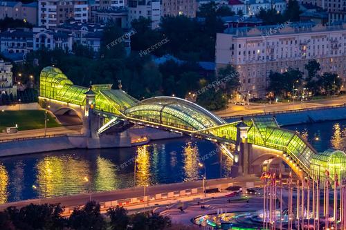 На фотографии запечатлен вид на Москву с высоты, где особое внимание привлекает Мост Богдана Хмельницкого, также известный как Киевский пешеходный мост. Это крытый пешеходный мост с подсветкой, который создает впечатляющий пейзаж вечерней Москвы. Фотография иллюстрирует современную городскую архитектуру и красоту ночного пейзажа. Ее можно использовать для городских туристических брошюр, путеводителей по Москве, рекламы туров и прочих материалов, привлекая внимание к туристическим достопримечательностям города.
