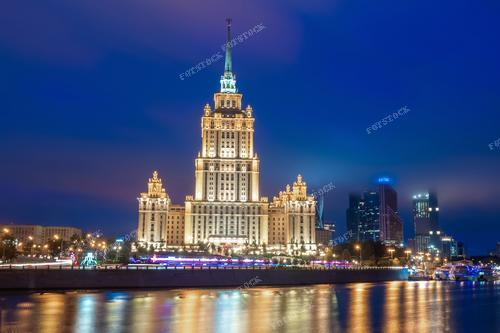 На фотографии изображено высотное здание на берегу Москвы-реки. Небоскреб выполнен в сталинском стиле архитектуры и запечатлен на фоне ночного неба. Фотография иллюстрирует характерный архитектурный стиль эпохи и узнаваемые здания столицы России.