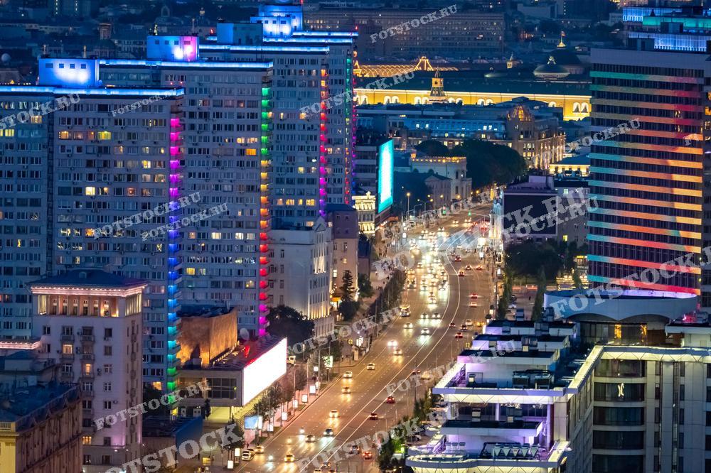 Фотография показывает ночной вид на улицы Москвы, где ярко освещенные здания и дороги создают красочную атмосферу. Видны многоэтажные дома с разноцветной подсветкой и оживленное движение автомобилей.