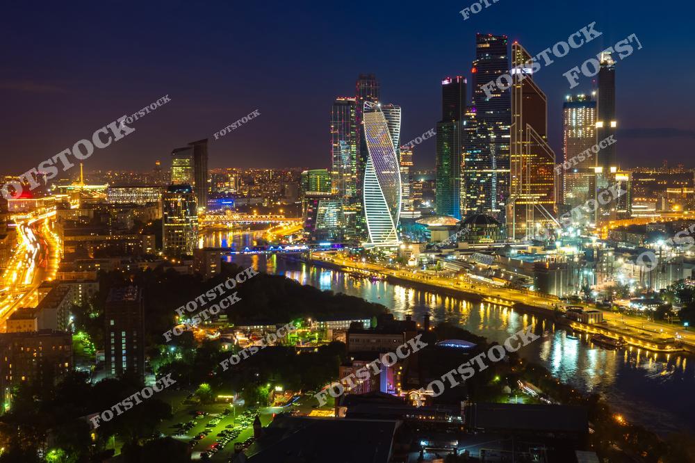 На фотографии изображен ночной вид на Москва-Сити, где небоскребы сверкают яркими огнями в темноте. Их отражение в реке Москве добавляет глубины и атмосферы снимку. Оживленные магистрали с потоком автомобилей подчеркивают энергетику ночной столицы. Этот вид представляет гармоничное сочетание современности и городской динамики, создавая впечатление мегаполиса, который никогда не спит.