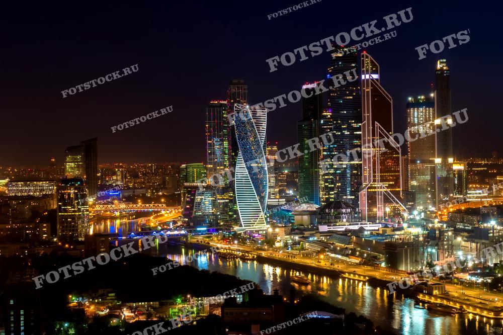 На фотографии изображена ночная панорама делового центра Москва-Сити. Яркие огни небоскребов отражаются в водах Москва-реки.