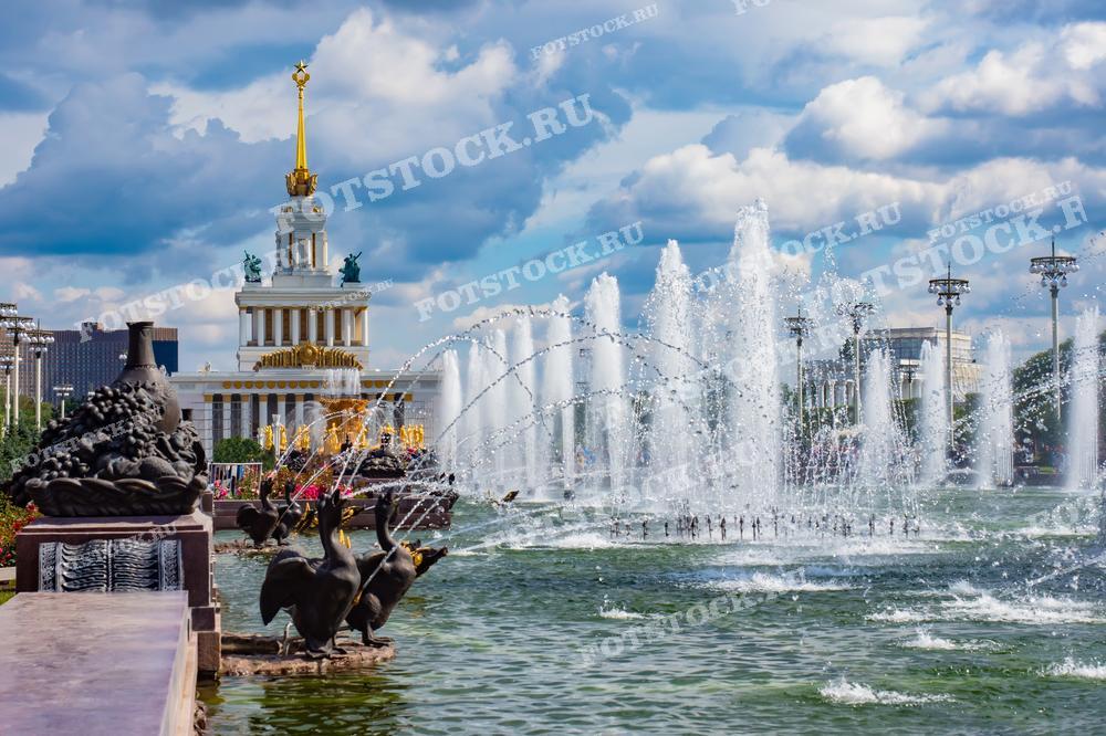 На фотографии изображены фонтан и Главный павильон ВДНХ в Москве. Вода из фонтана сверкает под лучами солнца, а на заднем плане виден величественный павильон со шпилем. Яркий летний день подчеркивает красоту этого места.