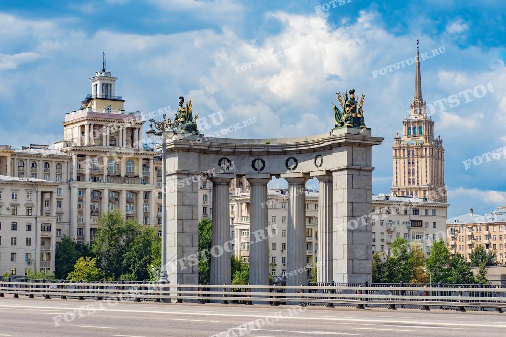 Фотография Триумфальной арки на Кутузовском проспекте в Москве с видом на сталинскую высотку и жилые дома в летний день. Подходит для использования в материалах о достопримечательностях Москвы, архитектуре и городской жизни.