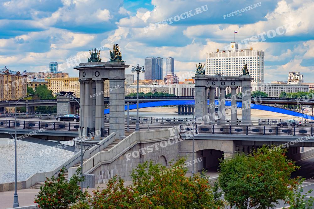 На фотографии изображен один из мостов Москвы с величественными арками и декоративными скульптурами. На заднем плане возвышается Дом Правительства Российской Федерации, также известный как Белый дом. Этот символический центр политической жизни России окружен современными зданиями и жилыми кварталами. Небо покрыто облаками, добавляя пейзажу драматический оттенок. Мост ведет через Москву-реку, и его архитектурные элементы придают городу монументальный и классический вид.