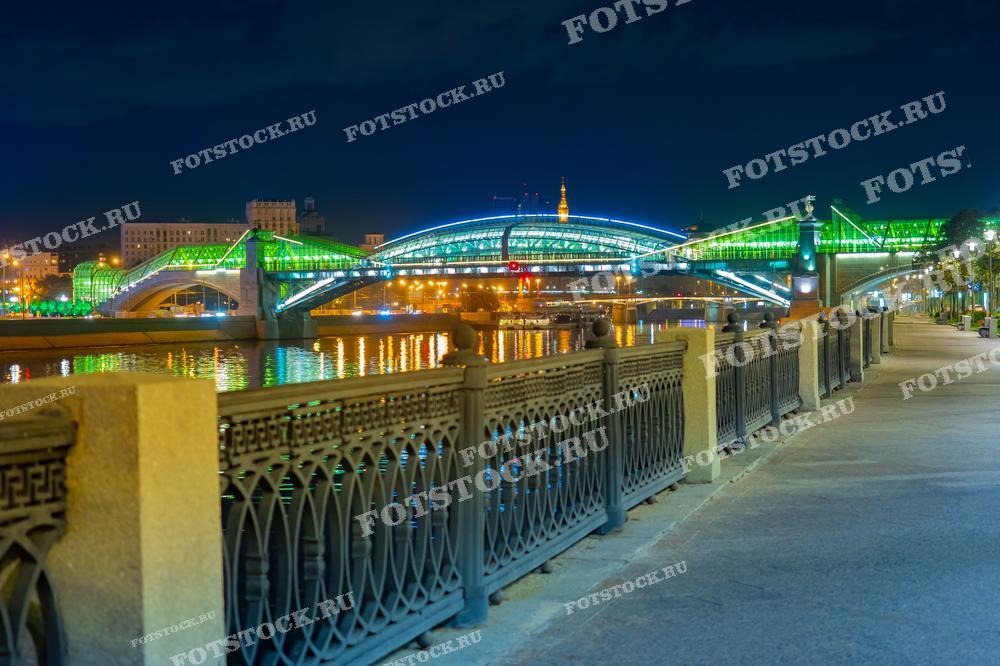На фотографии изображен мост Богдана Хмельницкого и набережная Москвы-реки в ночное время. Яркое освещение моста и его отражения в воде создают живописный ночной вид. Пустая набережная подчеркивает спокойную атмосферу ночного города.