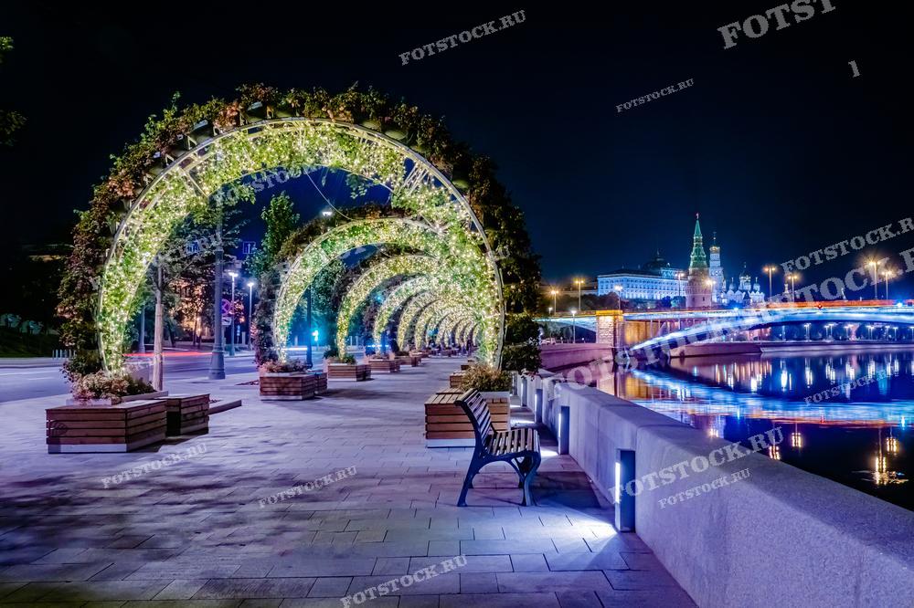 На этой фотографии изображена набережная Москвы, украшенная арками из цветов и гирлянд. Арки создают туннель света, направляющий взгляд к далёкому виду на Кремль и освещённый мост через Москва-реку. Такое оформление делает прогулку по набережной особенно романтичной и памятной. Это изображение идеально подойдёт для статей о ночной жизни города, о культурных и туристических привлекательностях Москвы.