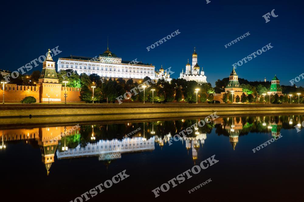 На фотографии изображен Московский Кремль в ночное время, освещенный яркими огнями. Красные стены Кремля и башни, включая знаменитую Спасскую башню, отражаются в спокойной воде Москвы-реки. На заднем плане виден Большой Кремлевский дворец и купола соборов. Вечернее освещение создает впечатляющий контраст между архитектурными элементами и темным небом, подчеркивая историческую и культурную значимость Кремля.