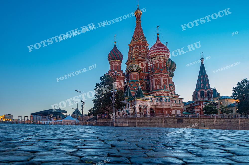 На фотографии изображен известный вид на собор Василия Блаженного в Москве, снятый с низкой точки у мощеной площади. Раннее утро или поздний вечер создают тихую и спокойную атмосферу, подчеркивая красочные купола и уникальную архитектуру собора. Такой ракурс и освещение подчеркивают величие и культурное наследие этого исторического памятника.