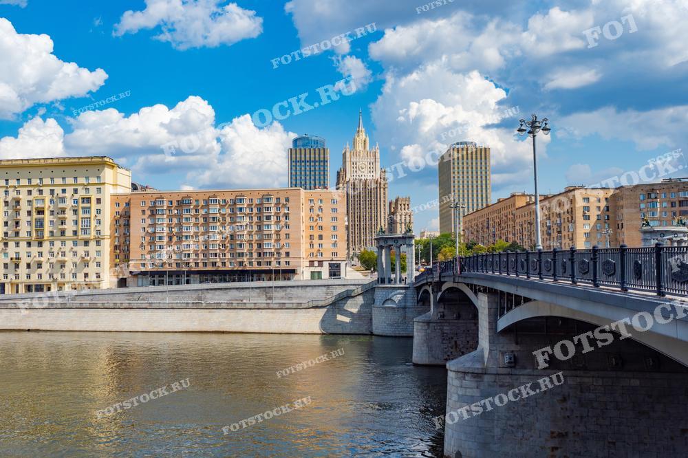 На фотографии изображен Бородинский мост через Москву-реку в летний день. На заднем плане виднеется здание Министерства иностранных дел России, одна из знаменитых сталинских высоток. Вдоль набережной расположены жилые дома и административные здания.