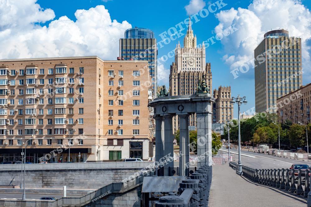 На фотографии изображен один из мостов Москвы с видом на здание Министерства иностранных дел РФ. Здание выделяется своим монументальным архитектурным стилем на фоне других построек. Справа и слева расположены современные здания и жилые дома, представляющие более поздние архитектурные эпохи. Голубое небо с облаками делает снимок светлым и живописным. Этот уголок Москвы сочетает в себе историю и динамику современного города.