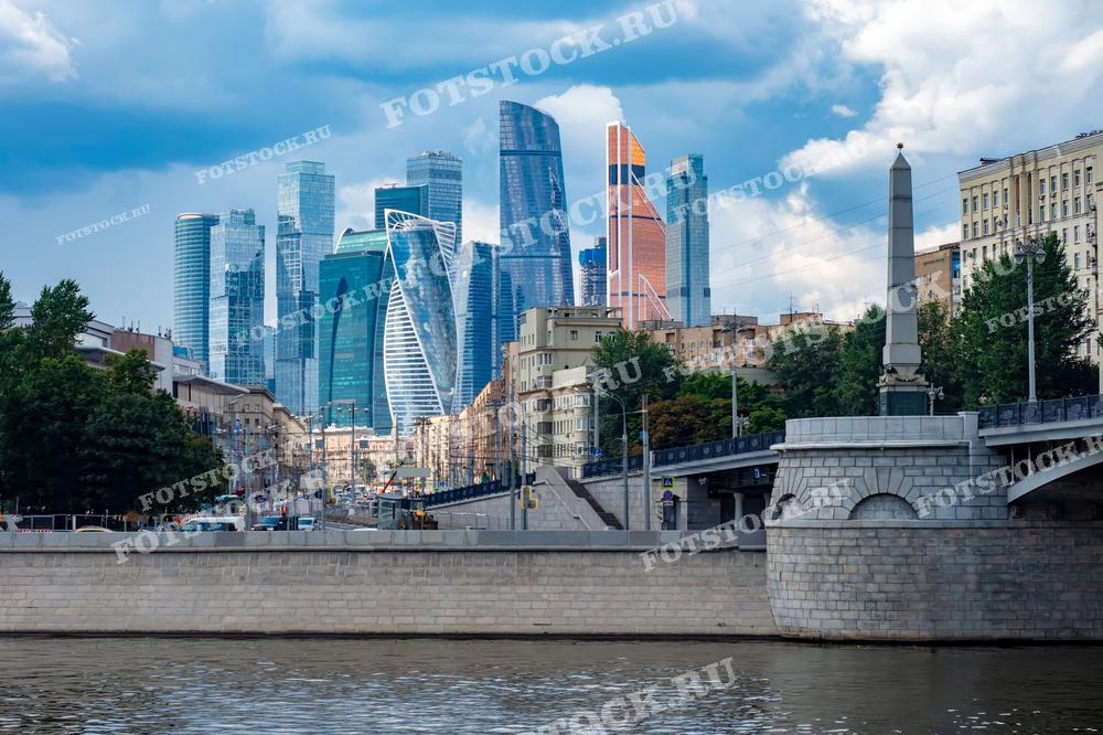 На фотографии изображен мост через Москву-реку на фоне небоскребов Москва-сити. В центре кадра видны современные офисные здания, отражающие динамичное развитие города. Вдоль набережной расположены жилые дома и административные здания.