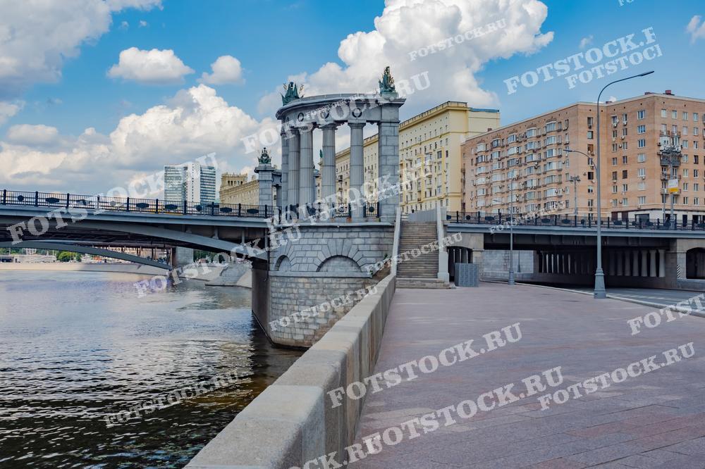 Фотография запечатлела один из мостов через Москву-реку, который соединяет два берега столицы. Мост украшен классическими колоннами и скульптурами, что придает ему особое архитектурное величие. На фоне видны типичные жилые дома Москвы, создающие атмосферу городской жизни. Тихие воды Москвы-реки и набережная подчеркивают спокойствие летнего дня. Мост является важной частью транспортной инфраструктуры города и одновременно украшением исторического центра.