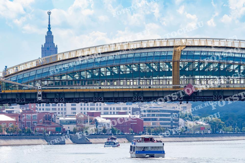 На фотографии изображен стеклянный пешеходный мост Богдана Хмельницкого, который соединяет два берега Москвы-реки. На заднем плане виднеется главный корпус Московского государственного университета (МГУ), одно из знаковых сталинских высотных зданий. По реке проходят прогулочные катера, добавляя динамики городской жизни.
