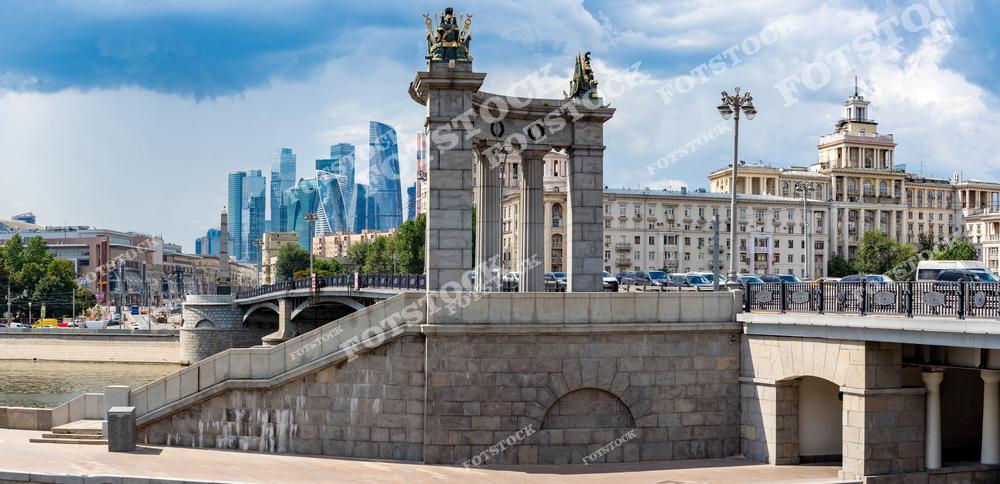 На фото изображена панорама Бородинского моста, который соединяет берега Москвы-реки. В кадре сочетаются здания, отражающие архитектуру Москвы разных эпох, от исторических сооружений до современных объектов. Летняя набережная Москвы-реки с её зелеными зонами и живописными видами является популярным местом для прогулок. Каменный мост, одна из значимых достопримечательностей столицы, выглядит величественно на фоне столицы, демонстрируя гармонию старинной архитектуры и современных городских пейзажей.
