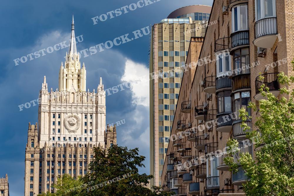 На фотографии изображены здание Министерства иностранных дел России и жилой дом на фоне голубого неба с облаками. Фото можно использовать для иллюстраций, рекламы и туристических материалов.