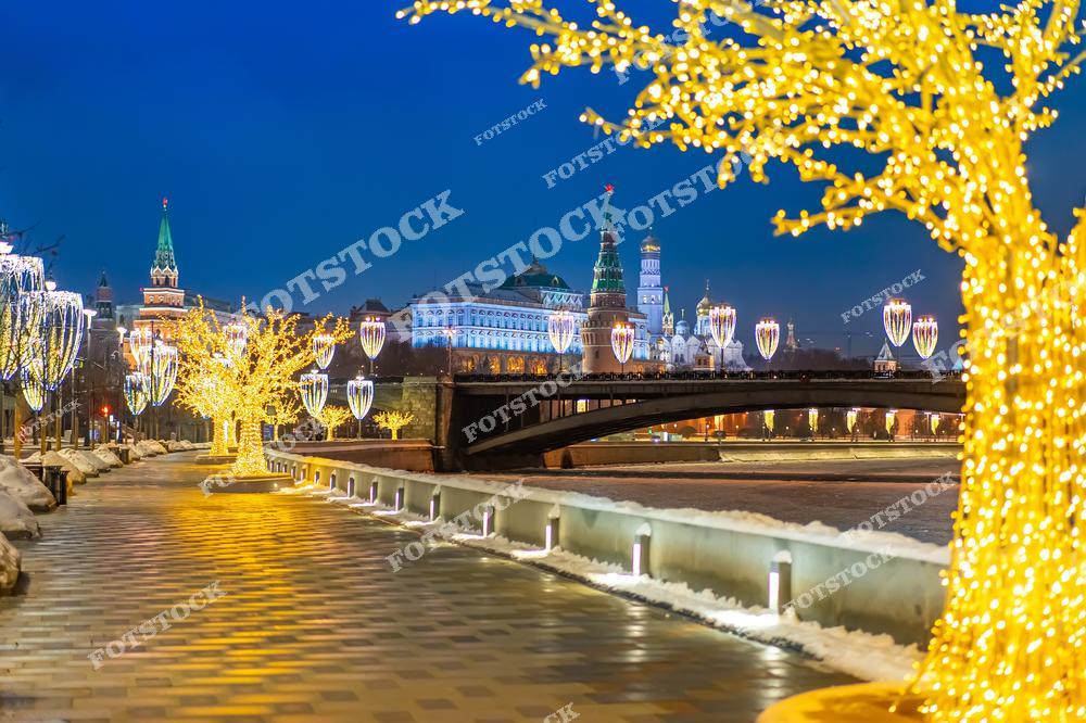 На фотографии запечатлена набережная Москвы-реки в зимний вечер, украшенная новогодней иллюминацией. Светящиеся деревья и декоративные элементы освещают дорожку вдоль воды. На заднем плане виден Большой Каменный мост, за которым возвышаются башни Кремля и колокольня Ивана Великого. Всё пространство оформлено празднично: световые украшения подчёркивают архитектуру и создают атмосферу волшебства. Отражения огней на воде усиливают эффект праздничного сияния. Москва предстает торжественной и уютной в зимнюю ночь.