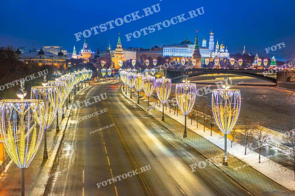 На фотографии изображен ночной вид на Кремль, одну из главных достопримечательностей Москвы, в зимний праздничный период. Улица вдоль Москвы-реки украшена яркими световыми декорациями в виде огромных гирлянд, которые создают ощущение праздника. Вдали видны величественные башни Кремля, а река частично покрыта льдом. Фотография демонстрирует зимнюю Москву в новогоднем убранстве, где улицы и здания сияют огнями, создавая атмосферу волшебства.