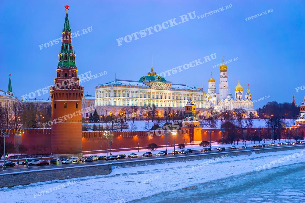 На фотографии изображен Московский Кремль зимним вечером. Кремлевские башни и Большой Кремлевский дворец красиво подсвечены. На переднем плане видна замерзшая Москва-река, вдоль которой движется автомобильный транспорт.