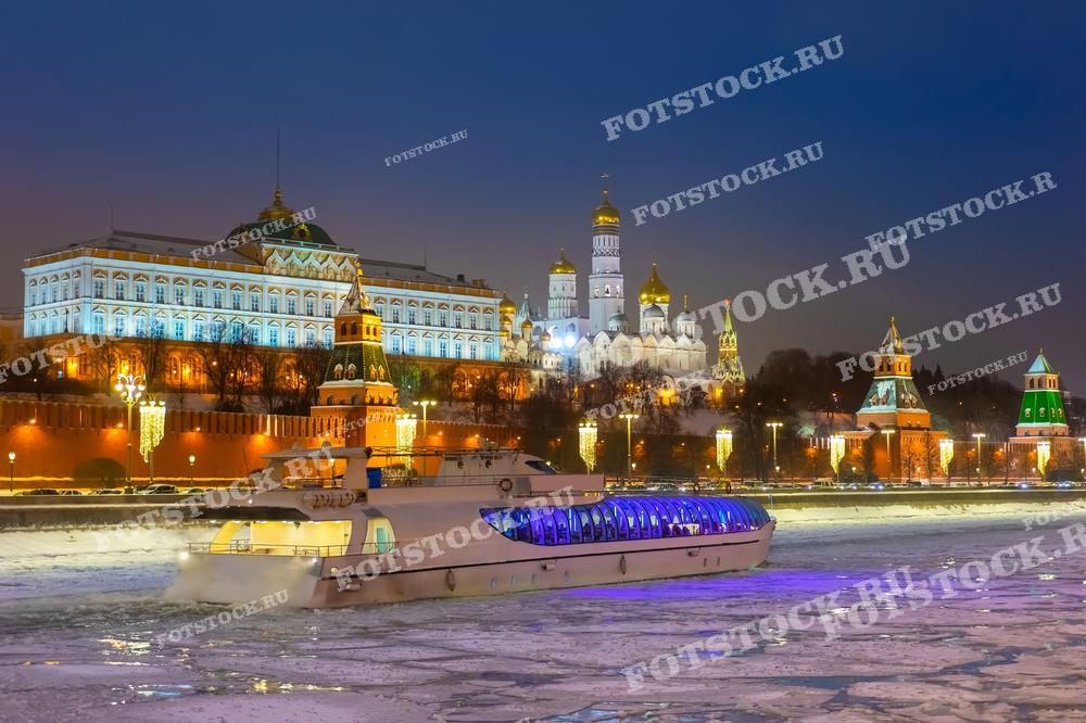 На фотографии представлен зимний вечерний вид на Московский Кремль и замерзшую Москву-реку. На переднем плане изображен прогулочный катер с яркой подсветкой, плавающий по ледяной реке, что добавляет красочности к уже живописному пейзажу. Эта фотография может использоваться в материалах, посвященных зимнему туризму в Москве, культурном наследии России или как часть декоративного оформления в публикациях о путешествиях.