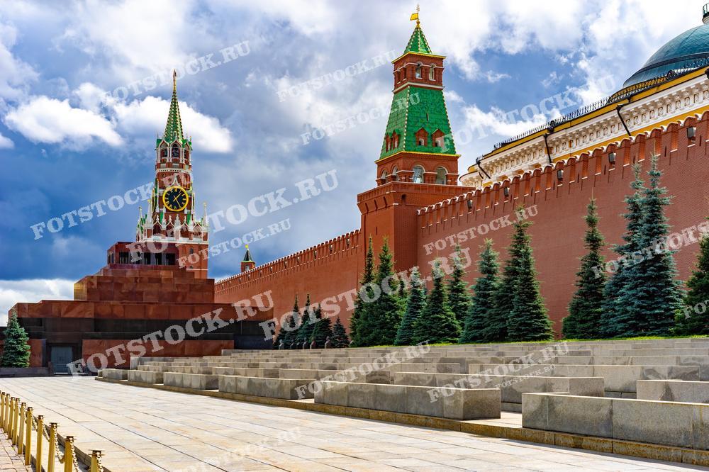 На фотографии изображена Красная площадь с видом на Мавзолей Ленина и Кремлевскую стену. В центре внимания — Спасская башня с знаменитыми курантами, которые являются символом времени и истории России. Мавзолей Ленина, построенный в 1930 году, расположен у подножия стены и является важной исторической достопримечательностью. Красная стена Кремля и стройные ряды елей добавляют величия архитектурному ансамблю. Фотография сделана в дневное время, когда над Москвой сгущаются облака.
