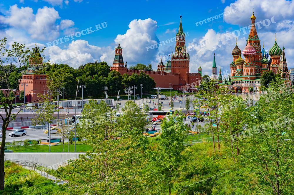 На фотографии изображён центр Москвы с территории парка Зарядье. На переднем плане можно увидеть зелёные насаждения парка, которые гармонично сочетаются с исторической архитектурой Кремля и Красной площади. Спасская башня Кремля с часами возвышается над красными стенами, а справа виднеется яркий Собор Василия Блаженного с его красочными куполами. Ясное небо и лёгкие облака придают фотографии атмосферу тёплого летнего дня.