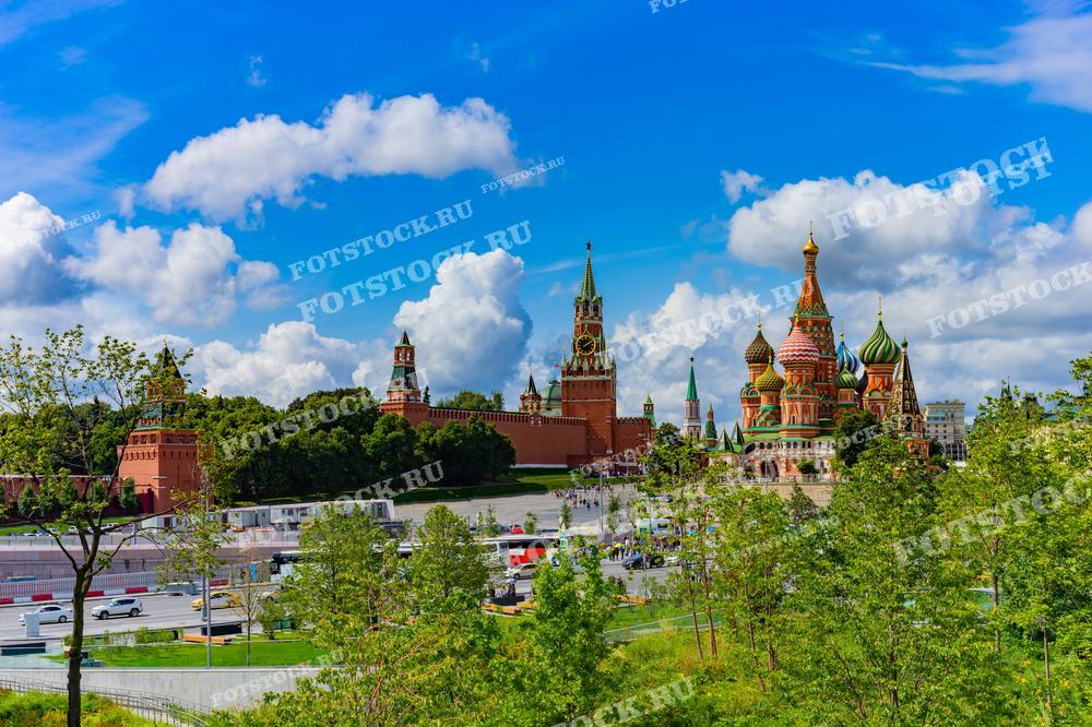 На фотографии изображён летний вид на главные достопримечательности Москвы: Кремль и Собор Василия Блаженного, сделанный из парка Зарядье. Спасская башня Кремля с часами доминирует над красными кремлёвскими стенами, создавая величественный фон. Собор Василия Блаженного с его разноцветными куполами располагается справа. Зелень парка Зарядье и прогулочные зоны гармонично вписываются в городской ландшафт.