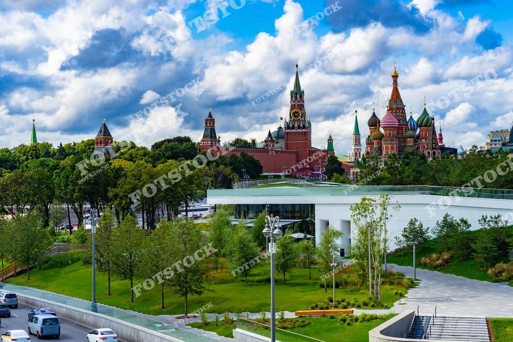 На фотографии изображен парк Зарядье в Москве, с которого открывается живописный вид на Кремль и Собор Василия Блаженного. В центре снимка видна Спасская башня Кремля с курантами, а справа – яркие купола собора. Зеленая зона парка с современным дизайном гармонично сочетается с исторической архитектурой. Голубое небо с белыми облаками добавляет свежести композиции. Этот уголок Москвы объединяет прошлое и настоящее, создавая уникальное место для прогулок.