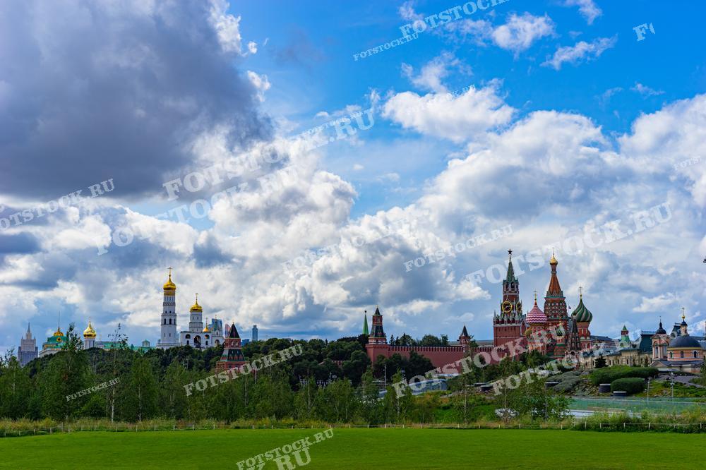 На фотографии изображены Кремль и Собор Василия Блаженного на Красной площади, видимые из Парка Зарядье в Москве. Яркий летний день подчеркивает зелень парка и облачное небо, создавая впечатляющий городской пейзаж.
