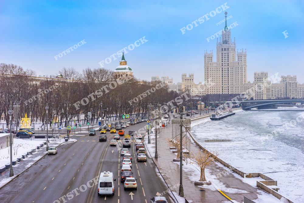 На фотографии изображена набережная Москвы-реки в зимний день. В центре кадра возвышается знаменитая сталинская высотка на Котельнической набережной, одна из самых узнаваемых достопримечательностей столицы. По дороге движется поток машин, подчеркивая оживленность города даже в морозное время года.