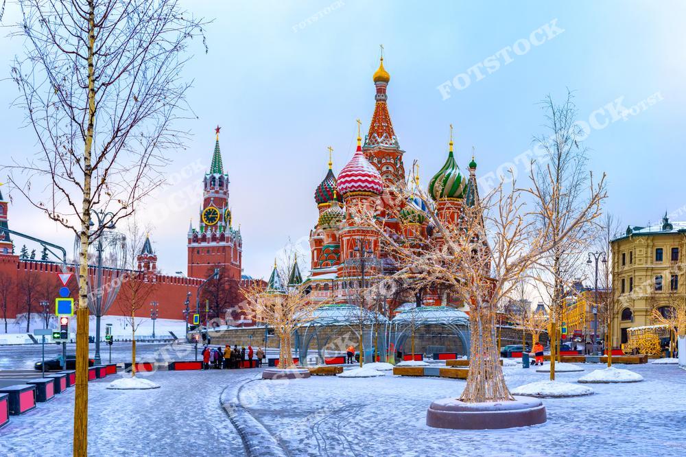 На фотографии изображен зимний пейзаж Красной площади в Москве. На переднем плане заснеженные деревья с подсветкой, добавляющие праздничности. Справа виден Собор Василия Блаженного с его яркими куполами, а слева — Спасская башня Кремля с часами. Снег и мягкий утренний свет придают композиции спокойствие и торжественность. Это место является не только символом столицы, но и центром притяжения туристов в зимний период.
