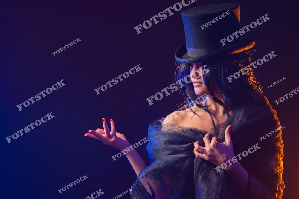 Фотография женщины в черном наряде и цилиндре на темном фоне с акцентным освещением. Позирование и игра света создают мистическую и таинственную атмосферу. Образ наполнен театральностью, элегантностью и загадочностью.