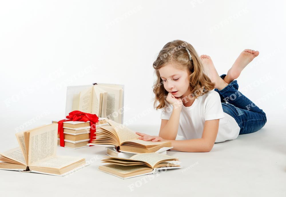 Фотография девочки в белой футболке и джинсах, лежащей на полу с книгой в руках. Вокруг нее разложены открытые книги и аккуратно сложенные стопки книг с красной лентой. Образ символизирует любовь к чтению, обучение и развитие ребенка.