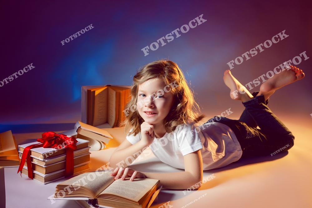 Фотография девочки с длинными светлыми волосами, лежащей на полу с книгой в руках. Вокруг нее расположены книги, перевязанные красной лентой и открытые страницы. Теплый свет и фон создают атмосферу уюта и сосредоточенности.