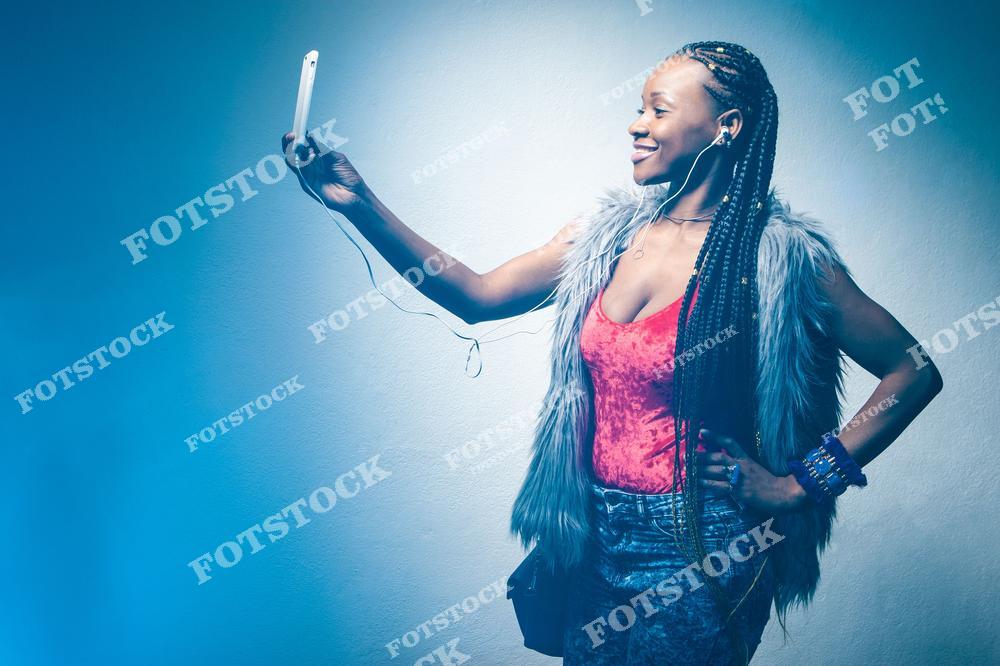Фотоснимок показывает афроамериканскую fashion-модель с африканскими косами, которая слушает музыку на телефоне и радостно улыбается, где изображение передаёт лёгкость, стиль и позитивное настроение. Эта фотография выглядит как современная иллюстрация городской жизни, а картинка создаёт яркий и динамичный слайд образа молодой африканской женщины с смартфоном.
