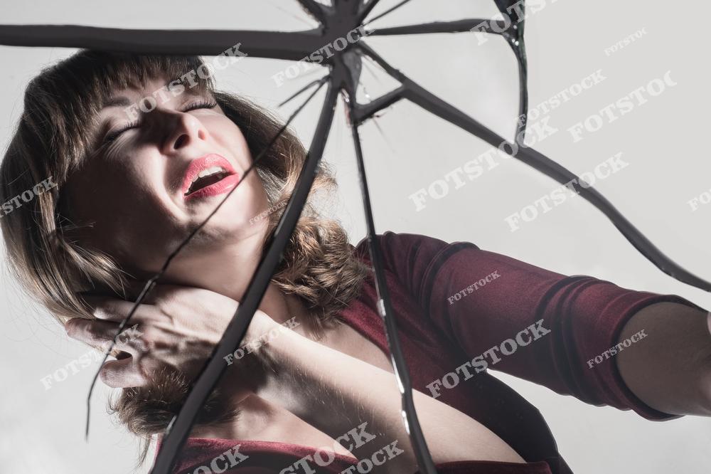 На фотографии женщина с закрытыми глазами и болезненным выражением лица смотрит сквозь трещины разбитого зеркала. Свет и композиция подчеркивают драматизм сцены. Этот образ символизирует внутреннюю борьбу, уязвимость и эмоциональный надлом.