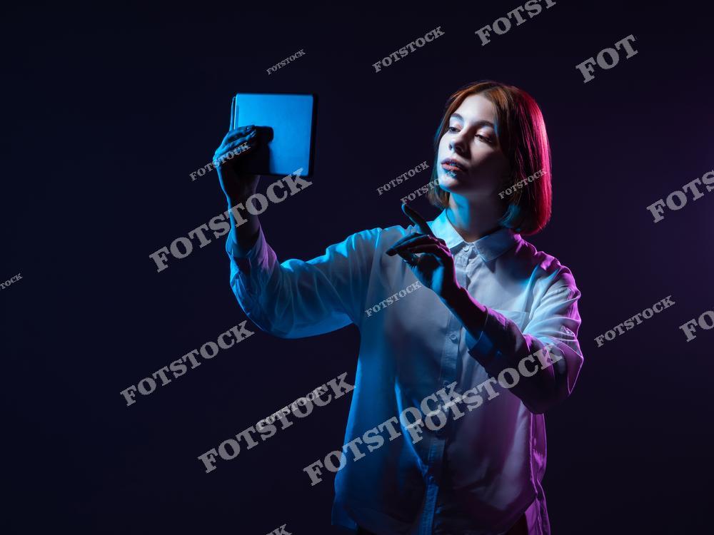 На фотографии изображена молодая женщина с планшетом, работающая с виртуальным интерфейсом. Темный фон с неоновыми оттенками синего и розового создает футуристическую атмосферу, символизируя цифровое будущее и инновации.